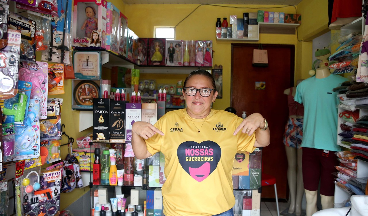 Rejane posa dentro da sua loja usando a camisa do programa nossas guerreiras